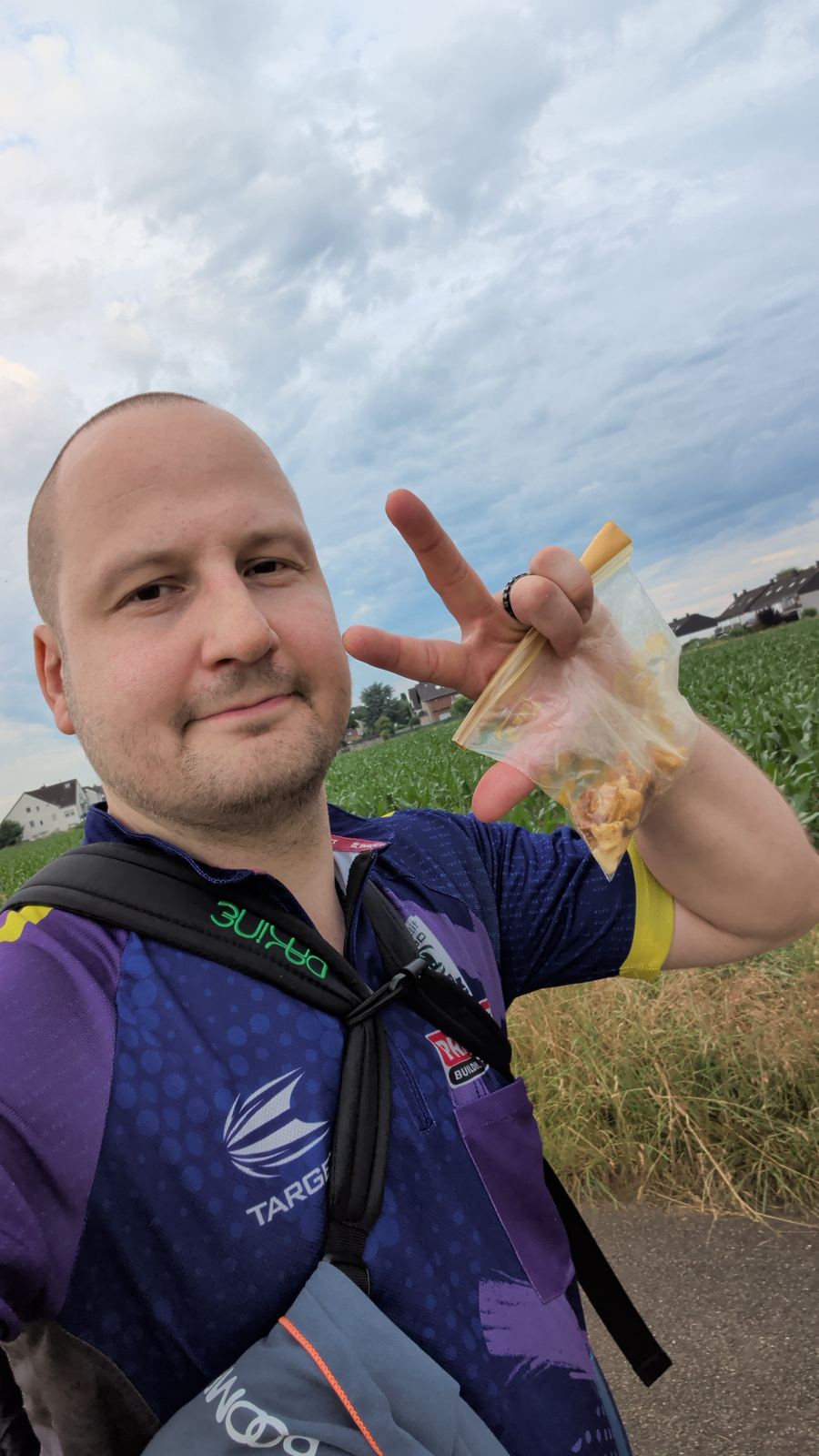 Selfie unterwegs: Rucksack mit Trinksystem, eine Hand hält eine kleine Tüte mit Proviant, im Hintergrund Felder unter bewölktem Himmel.