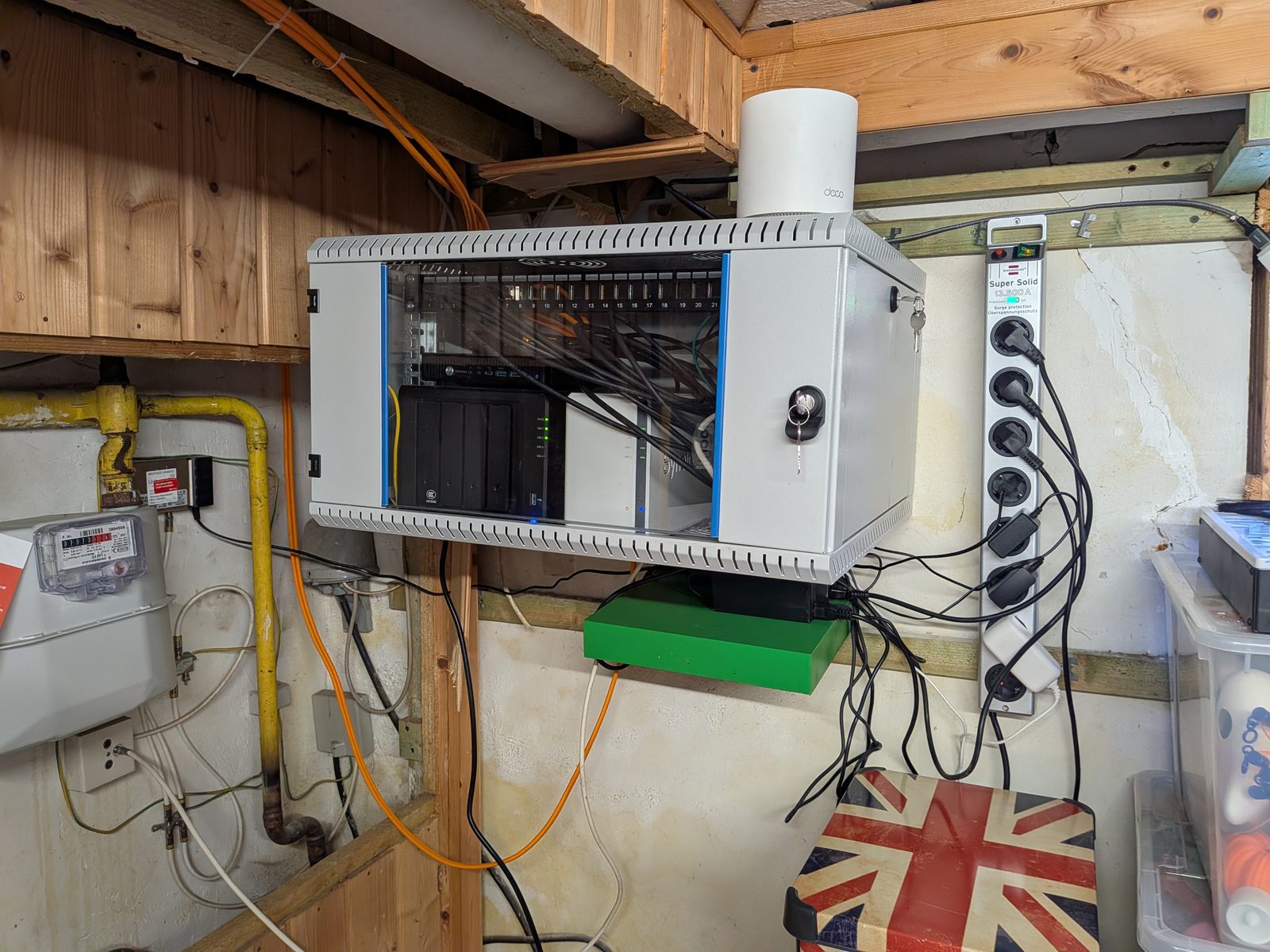 Der neue Serverschrank an der Kellerwand, Tür geöffnet: Switch, Patchfeld und NAS-Gehäuse sauber verkabelt, daneben eine Steckdosenleiste mit Überspannungsschutz.