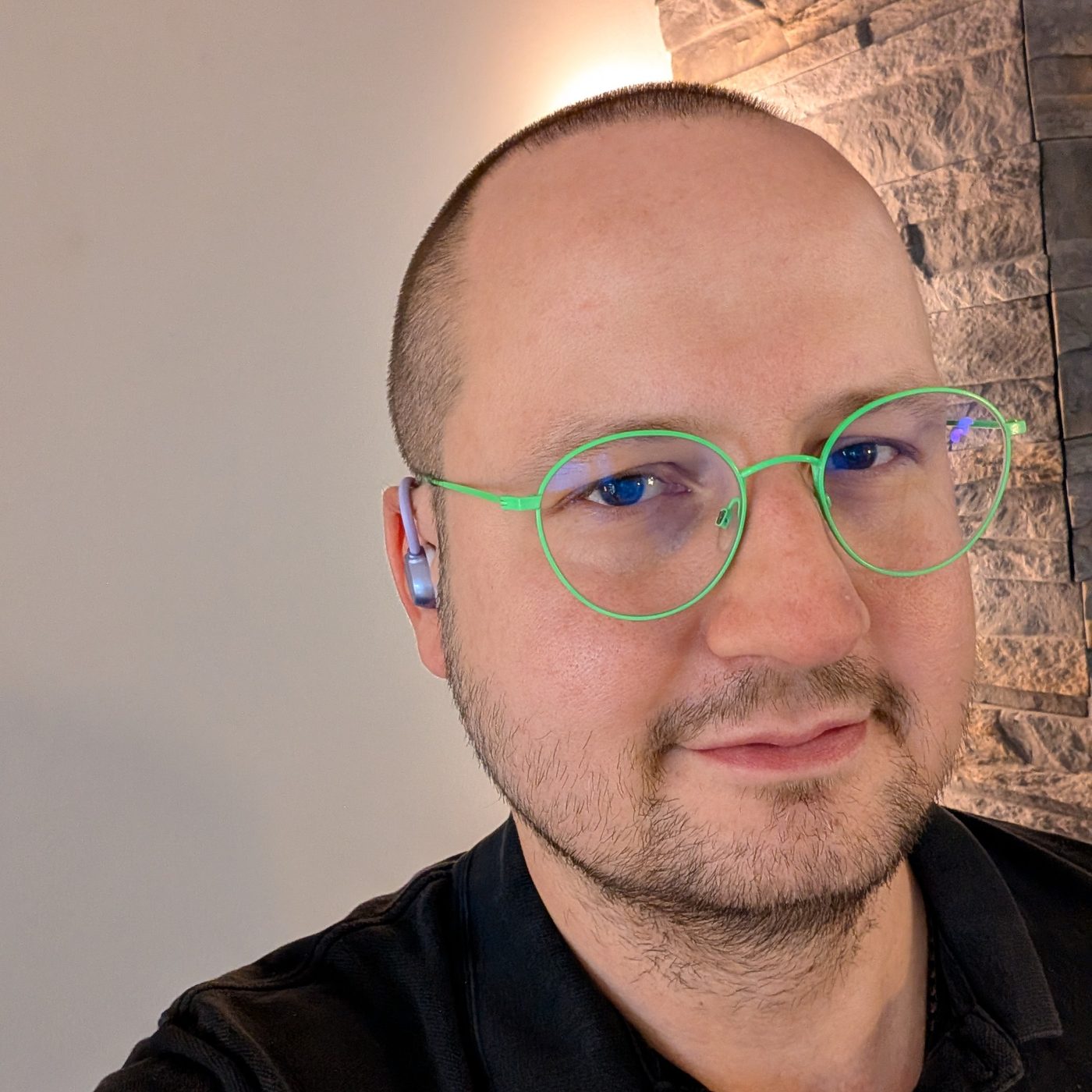 Selfie mit grüner Brille, im linken Ohr steckt ein grauer Bluetooth-Earbud