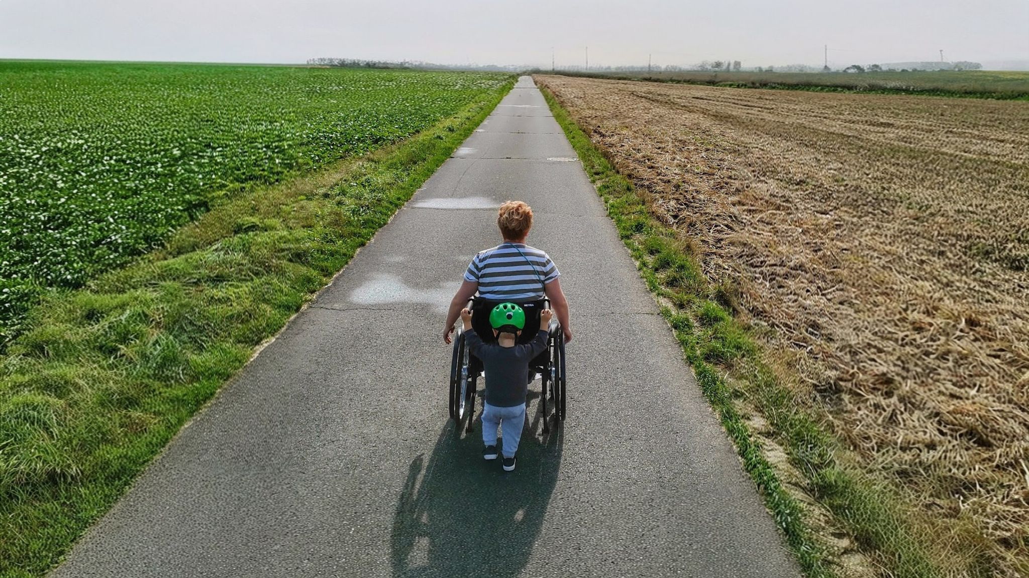 Aylin im Rollstuhl auf einem Feldweg, daneben unser Sohn mit grünem Helm