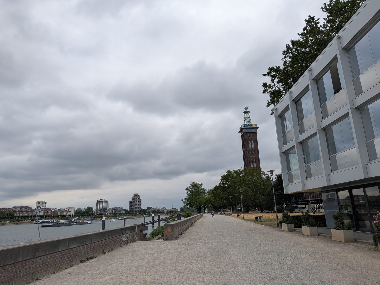 Blick über die Rheinpromenade in Köln-Deutz an einem bewölkten Tag: links der Rhein mit einem Frachtschiff und der Kölner Uferzeile, rechts ein modernes Bürogebäude, im Hintergrund der alte Messeturm mit dem RTL-Schriftzug.